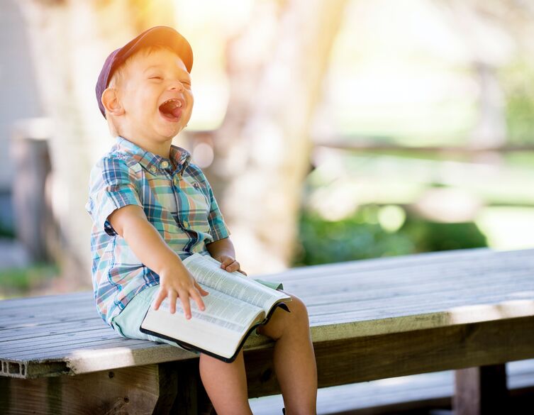 laughing boy wearing a cap and holding a book while sitting on a bench outside